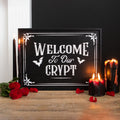 Black framed wooden wall sign with white gothic lettering reading Welcome To Our Crypt with bat and decorative corner details, styled with black and red candles and red roses