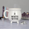White ceramic cauldron-shaped mug with White Witch text and crescent moon detail, styled with amethyst crystals, crystal towers and tealight candles