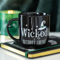 Black ceramic mug with white Wicked Without Coffee slogan and witch hat graphic, resting on a spell book with a green candle in the background