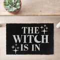 Black coir door mat with The Witch Is In slogan in white lettering with sparkle star details, styled on a wooden floor with a succulent plant nearby