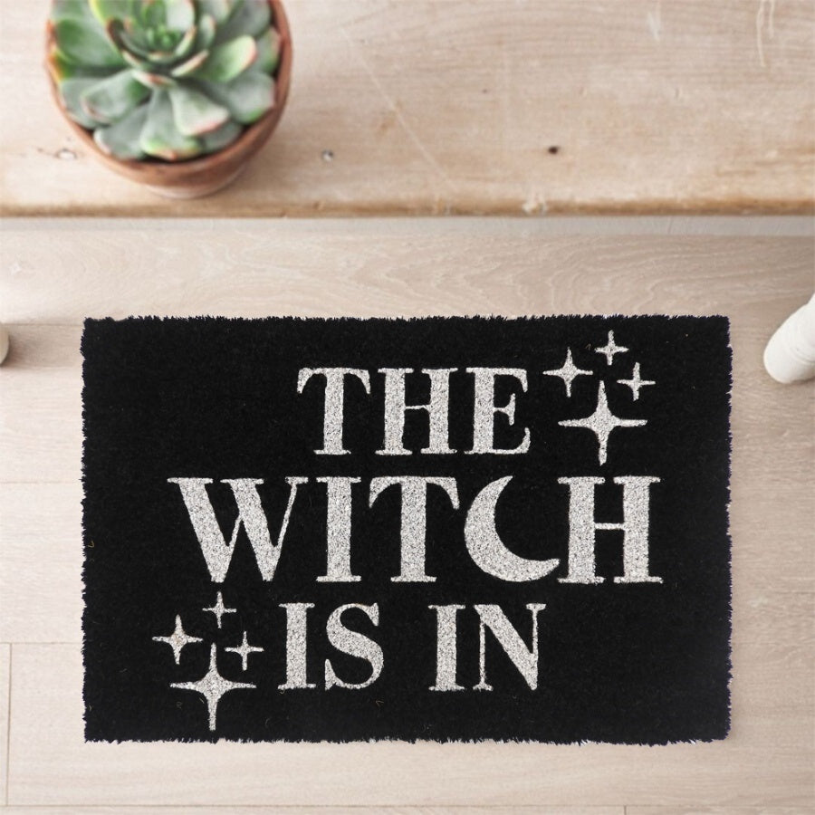 Black coir door mat with The Witch Is In slogan in white lettering with sparkle star details, styled on a wooden floor with a succulent plant nearby