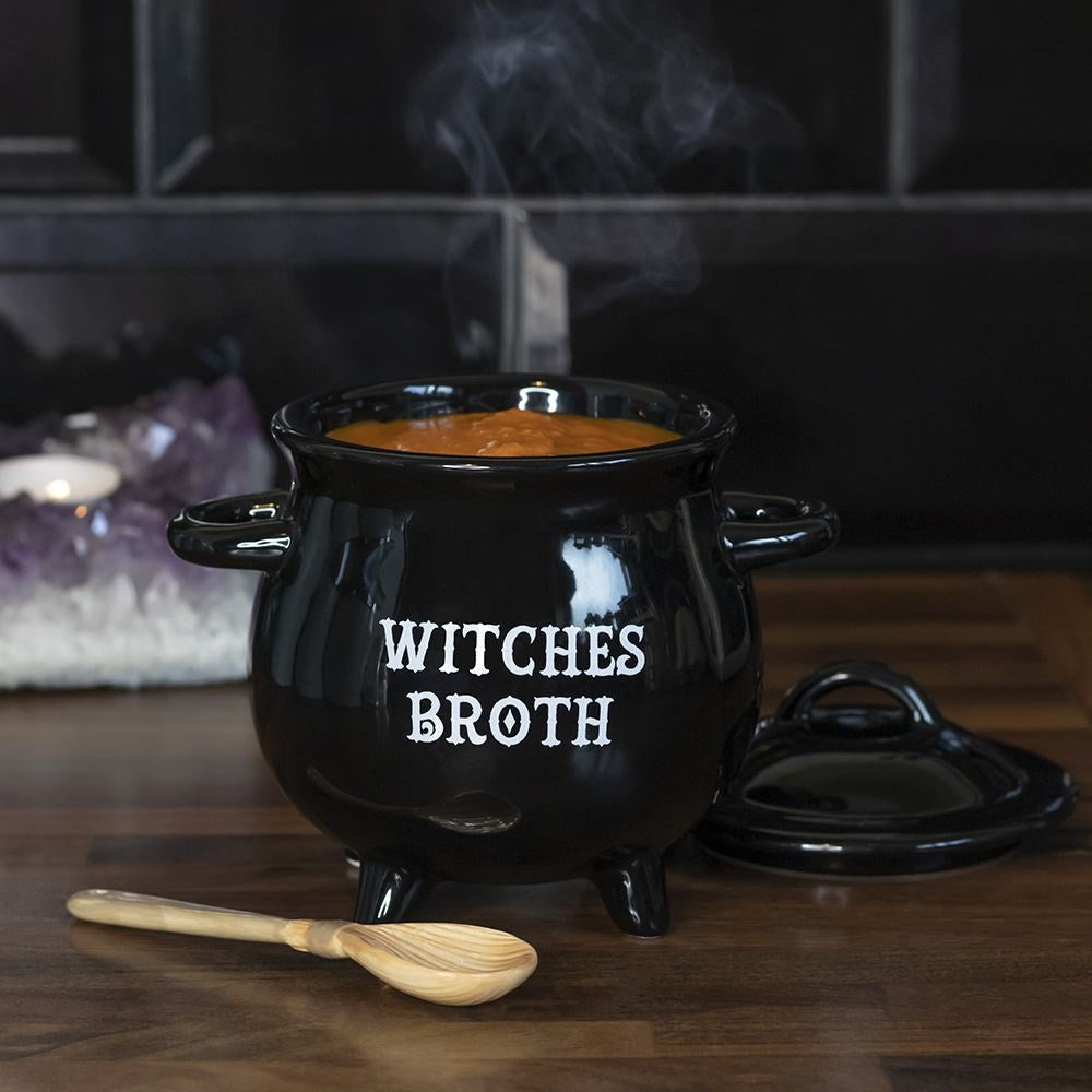 Black ceramic cauldron-shaped soup bowl with Witches Broth text and lid, filled with soup and styled on a dark wooden surface with a wooden broom spoon and amethyst crystals
