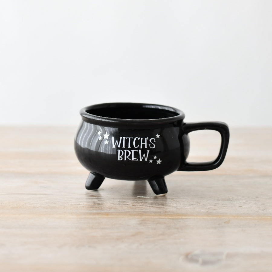 Black ceramic Witch's Brew cauldron mug with three legs, square handle and white star detailing, on a wooden surface