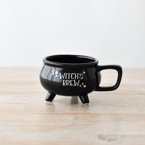 Black ceramic Witch's Brew cauldron mug with three legs, square handle and white star detailing, on a wooden surface