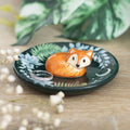 Dark green ceramic trinket dish with a 3D curled orange fox figurine and botanical fern leaf design, displayed on a wooden surface with a silver ring resting beside the fox and white gypsophila flowers in the foreground